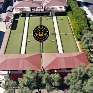 Aerial view of a sports complex featuring four pickleball courts marked with the gladiator logo, surrounded by red canopies and greenery.