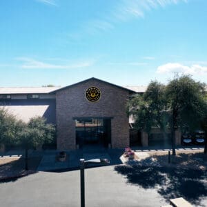 Aerial view of a single-story building with a circular logo above the entrance, surrounded by a parking lot and sparse vegetation under a clear sky.