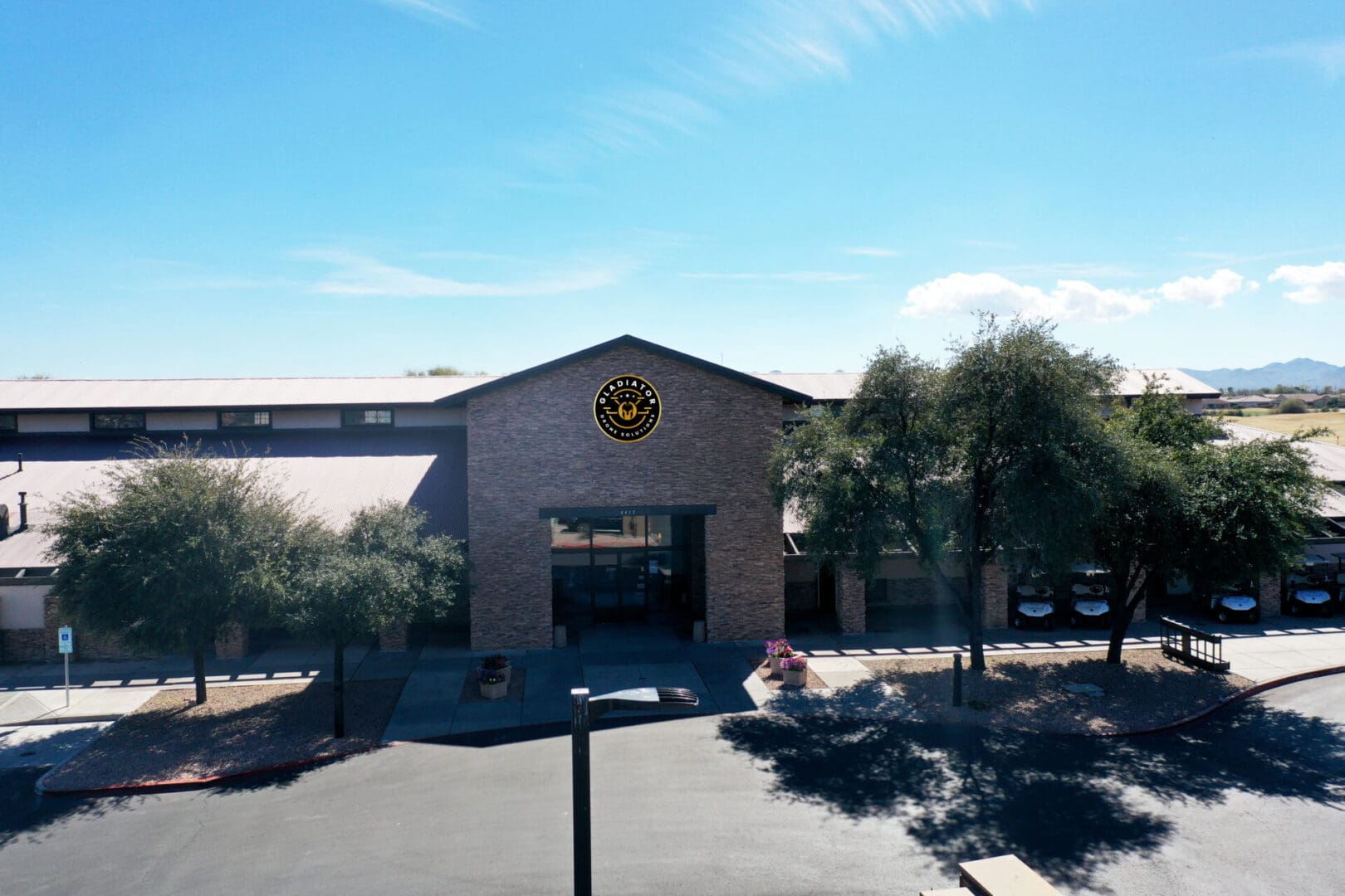 Aerial view of a single-story building with a circular logo above the entrance, surrounded by a parking lot and sparse vegetation under a clear sky.