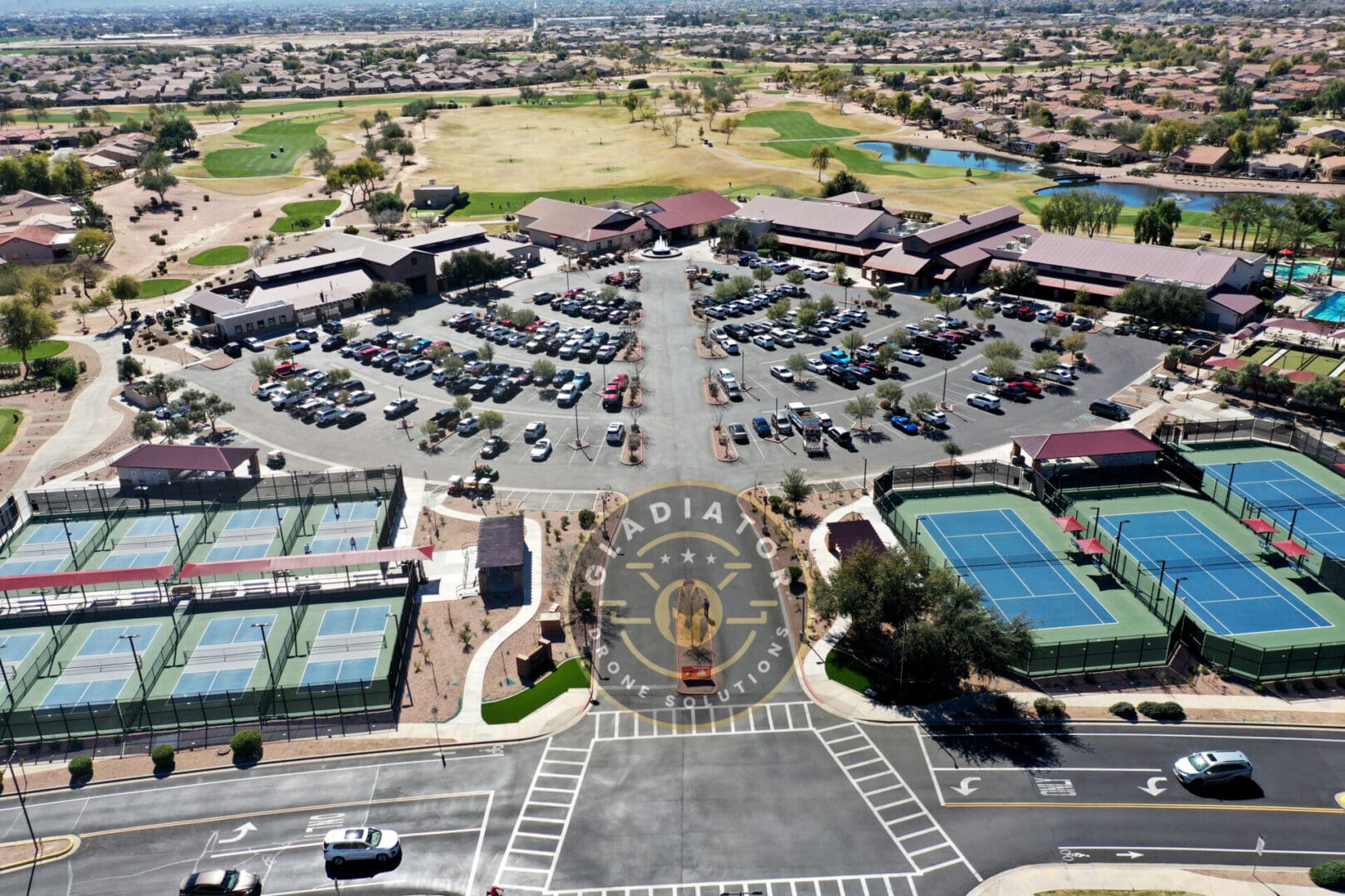 Aerial view of a large parking lot at a sports complex featuring tennis courts and golf courses, with surrounding residential areas.