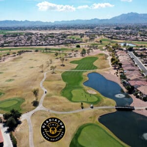 Aerial view of a golf course with green patches and water features, surrounded by residential houses, set against a mountainous backdrop.