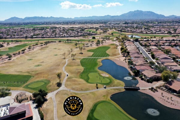 Aerial view of a golf course with green patches and water features, surrounded by residential houses, set against a mountainous backdrop.