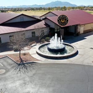 Aerial view of a large building with a distinctive, dark roof featuring a round fountain at the front, and a sign depicting a bear and the text "gold canyon." located in a flat, green landscape.