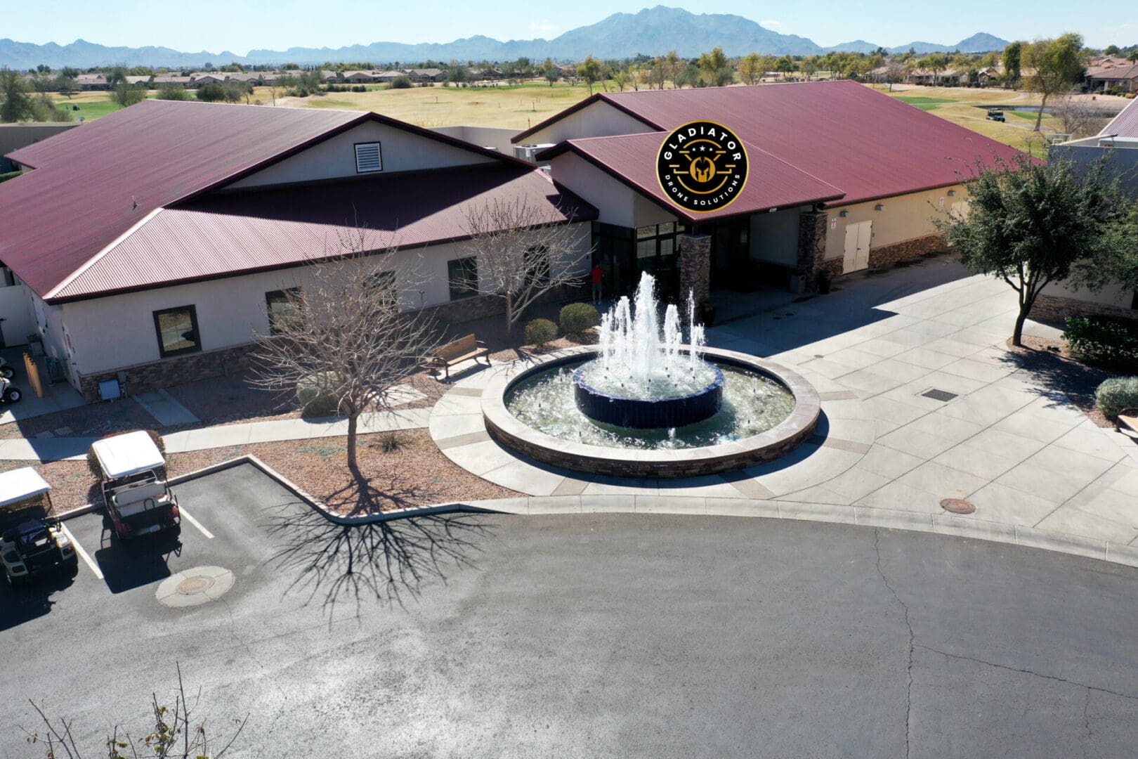 Aerial view of a large building with a distinctive, dark roof featuring a round fountain at the front, and a sign depicting a bear and the text "gold canyon." located in a flat, green landscape.