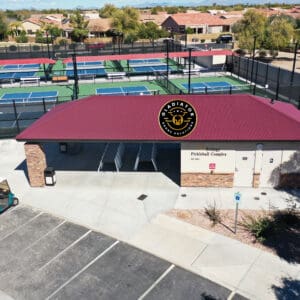 Aerial view of a single-story building labeled "pickleball complex" with a red roof, adjacent to several pickleball courts, in a sunny, suburban setting.