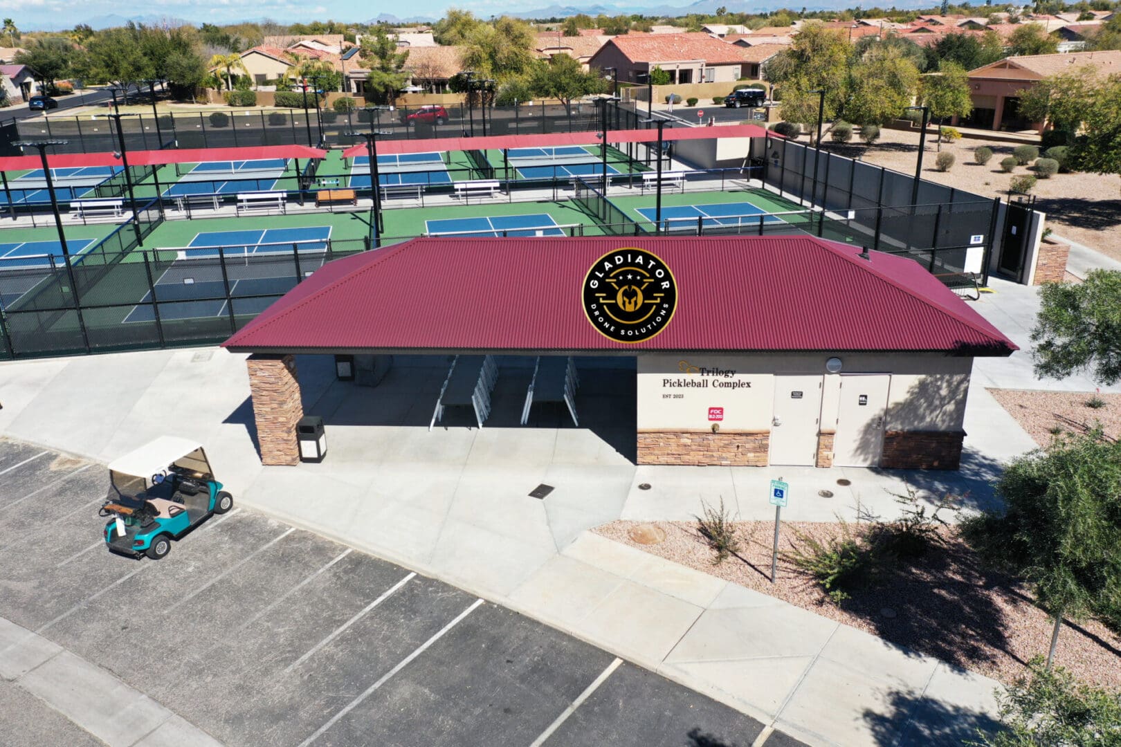 Aerial view of a single-story building labeled "pickleball complex" with a red roof, adjacent to several pickleball courts, in a sunny, suburban setting.