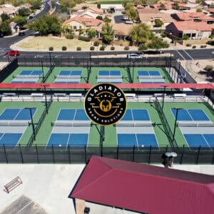 Aerial view of six green pickleball courts enclosed by black fences, with surrounding residential area and a parking lot.