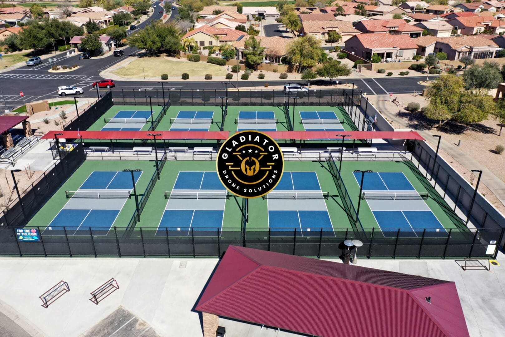 Aerial view of six green pickleball courts enclosed by black fences, with surrounding residential area and a parking lot.