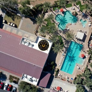 Aerial view of a hotel with two distinct swimming pools, surrounded by lounging areas, and adjacent red-roofed buildings.