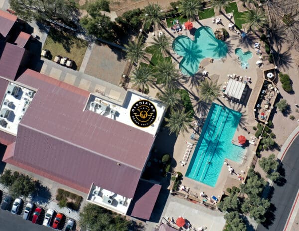 Aerial view of a hotel with two distinct swimming pools, surrounded by lounging areas, and adjacent red-roofed buildings.