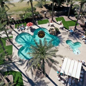 Aerial view of a resort with uniquely shaped swimming pools, sun loungers, and a dining area under umbrellas, surrounded by palm trees.