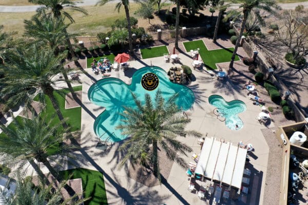 Aerial view of a resort with uniquely shaped swimming pools, sun loungers, and a dining area under umbrellas, surrounded by palm trees.