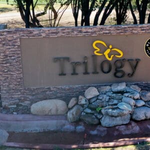 Aerial view of a stone entrance sign for trilogy with a logo, surrounded by rocks and greenery.