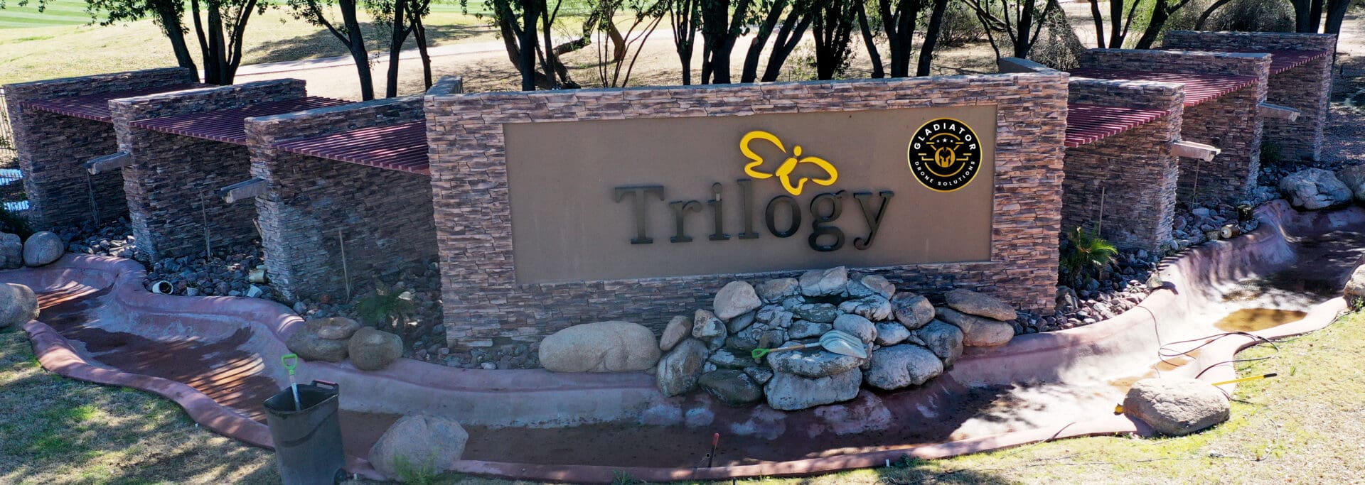 Aerial view of a stone entrance sign for trilogy with a logo, surrounded by rocks and greenery.