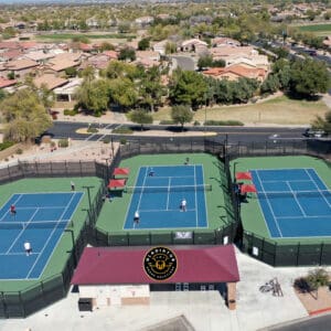 Aerial view of a tennis facility with four courts, players in action, surrounded by residential area, featuring a central clubhouse with logos.