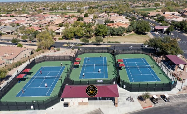 Aerial view of a tennis facility with four courts, players in action, surrounded by residential area, featuring a central clubhouse with logos.