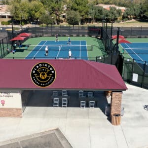 Aerial view of a tennis complex with multiple courts and players, featuring a building with a logo, bicycles parked outside, in a suburban setting.