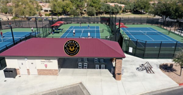 Aerial view of a tennis complex with multiple courts and players, featuring a building with a logo, bicycles parked outside, in a suburban setting.