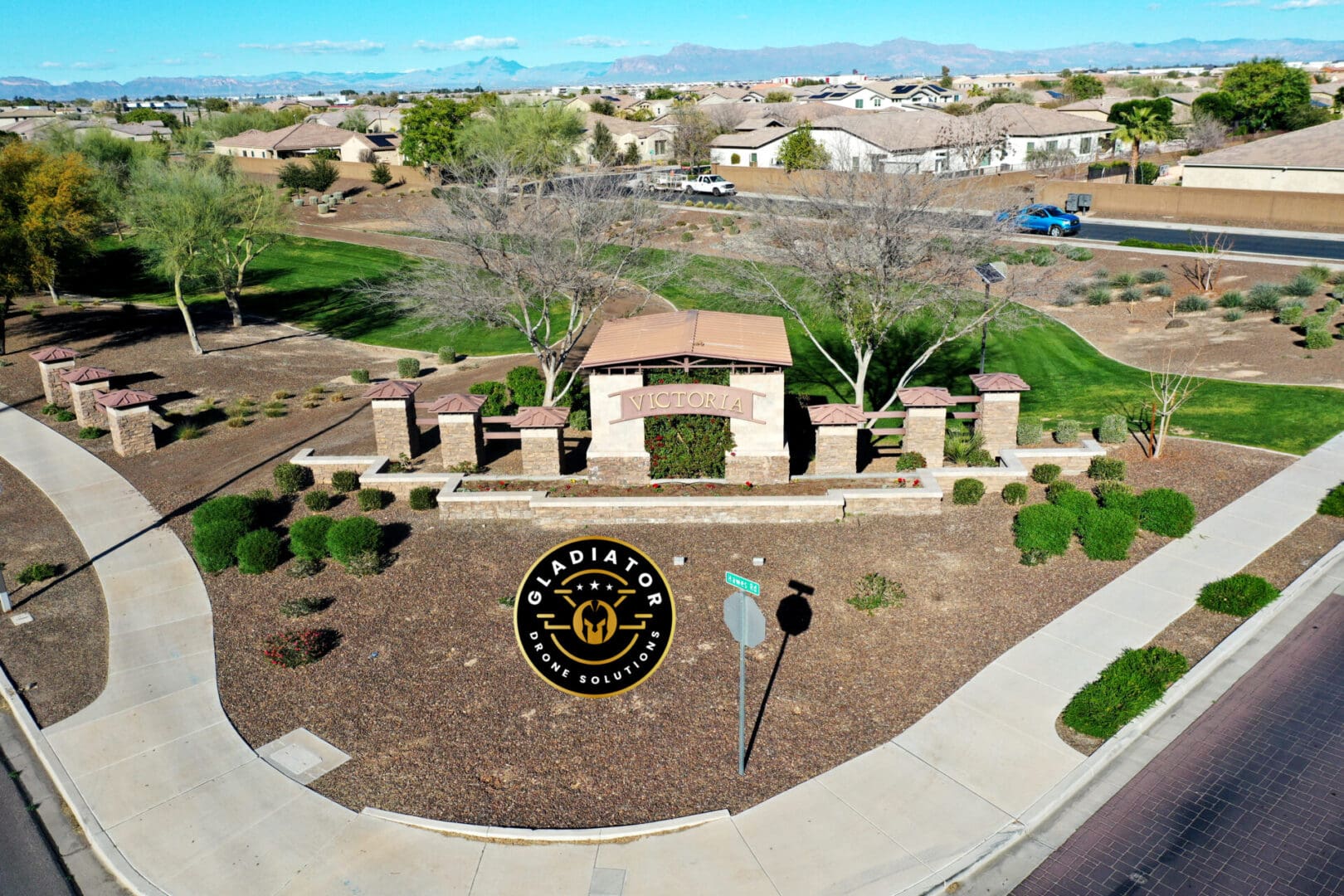 Aerial view of the entrance to a residential area called "victoria," featuring a landscaped roundabout with a decorative sign and seal, surrounded by homes and a clear blue sky.