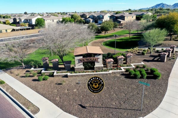 Aerial view of a suburban entrance sign reading "victoria" with surrounding neatly manicured lawns and residential homes, set against a mountain backdrop.