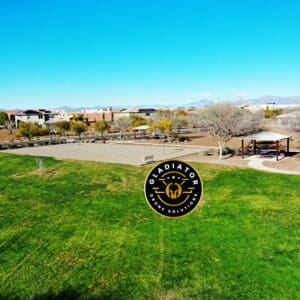 Aerial view of a suburban park with green fields, playground equipment, and houses in the background under a clear blue sky.