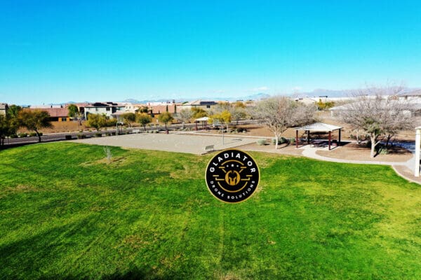 Aerial view of a suburban park with green fields, playground equipment, and houses in the background under a clear blue sky.