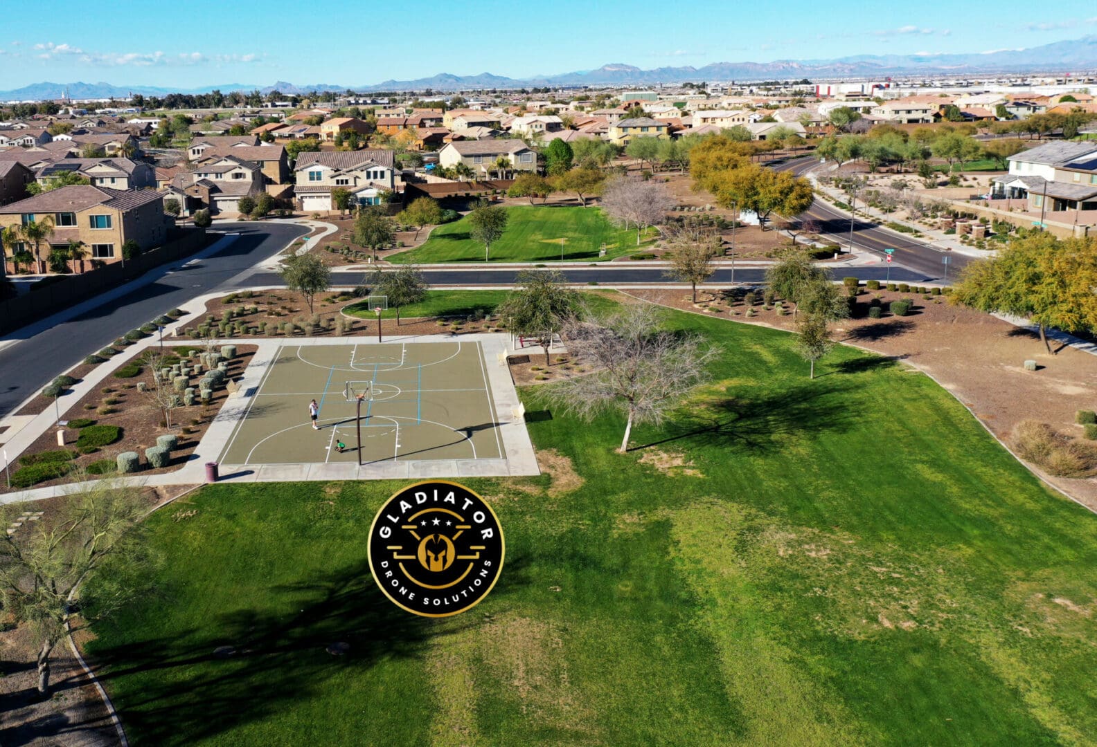 Aerial view of a suburban neighborhood featuring a basketball court and park, with houses and mountainous background.