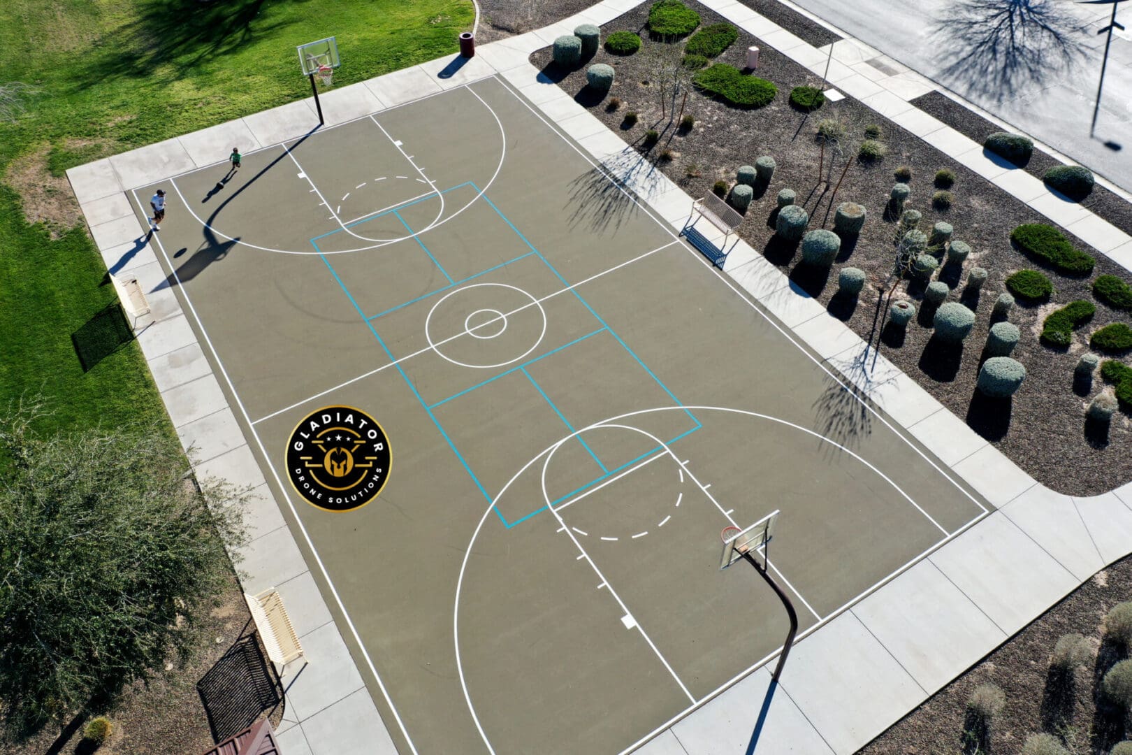 Aerial view of an outdoor basketball court with distinctive markings and a logo at the center, surrounded by landscaping and trees.