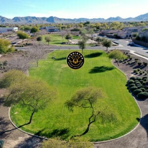 Aerial view of a suburban park with green grass and scattered trees, surrounded by houses, with mountains in the background and a logo superimposed in the center.