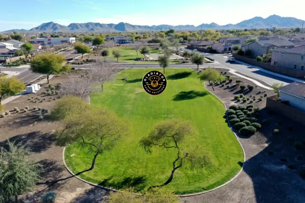 Aerial view of a suburban park with green grass and scattered trees, surrounded by houses, with mountains in the background and a logo superimposed in the center.