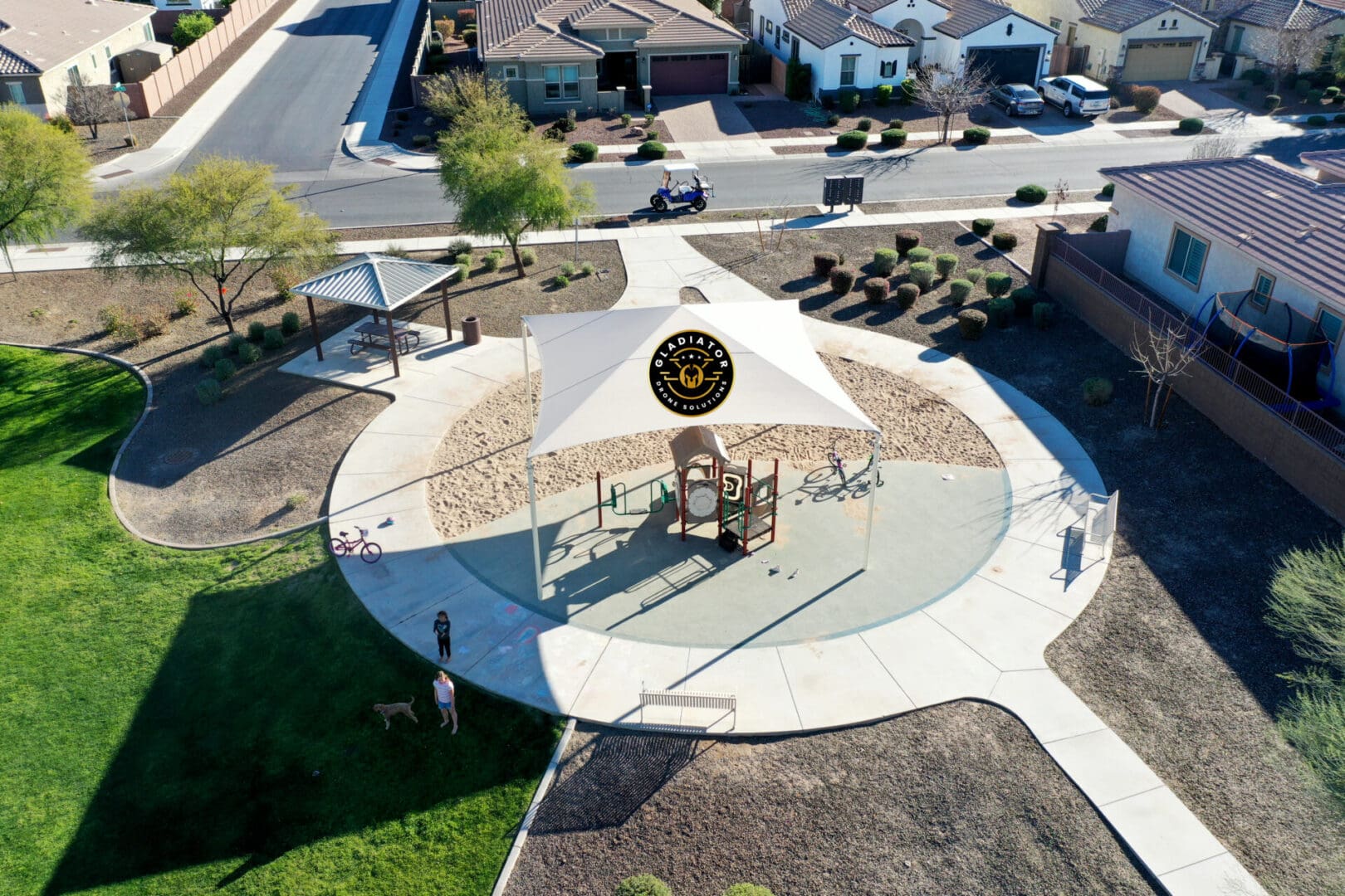 Aerial view of a suburban park with a circular playground, basketball court, and walking paths, surrounded by residential houses.