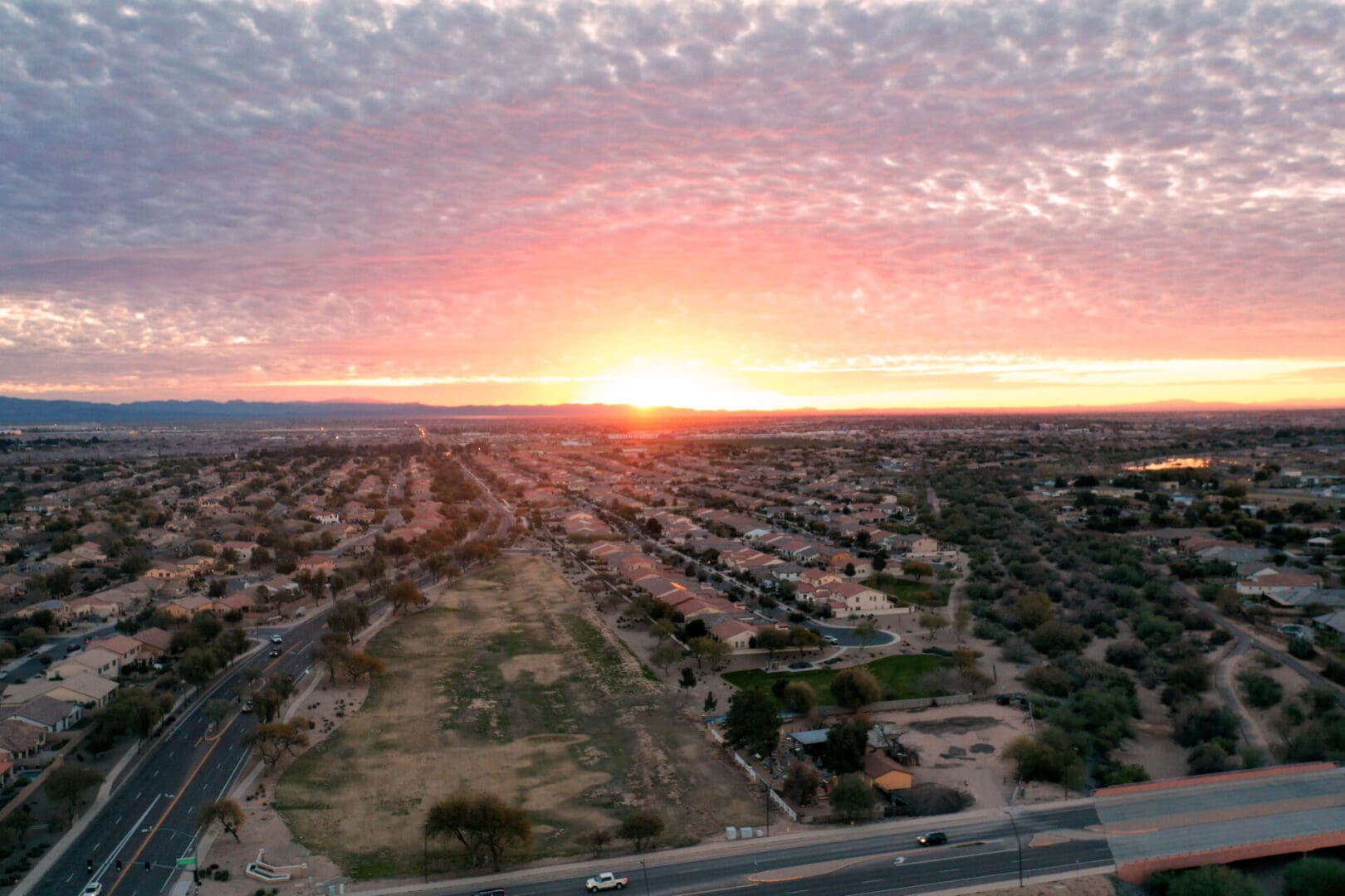 Aerial view of a suburban area at sunset, showcasing a vibrant sky with a pronounced orange glow and scattered clouds over rows of houses and a highway.